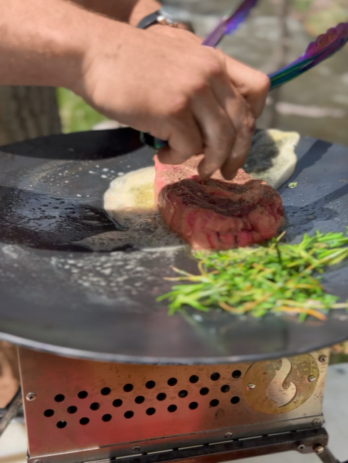 A hand places a piece of raw steak onto the hot LavaBox SWOK Discada Combo with melted butter, next to fresh rosemary.