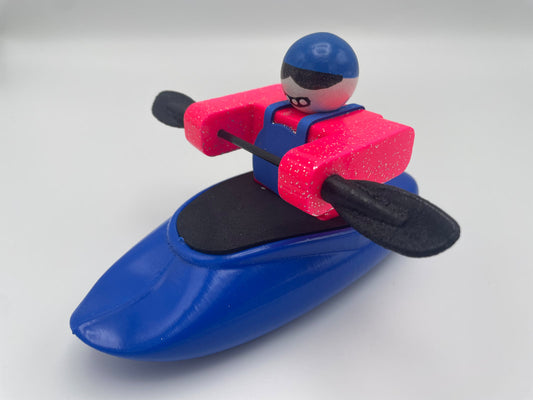 A toy figure from the Foamie Friends collection, known as the Foamie Spud Play Boat, features a person kayaking in a blue kayak. They are wearing a blue helmet and a pink vest while holding black paddles as they navigate river dynamics.
