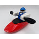 A Foamie Friends character in a blue outfit and helmet is seated in a Foamie Spud Play Boat, holding a black paddle against a white background, ready to explore river dynamics.