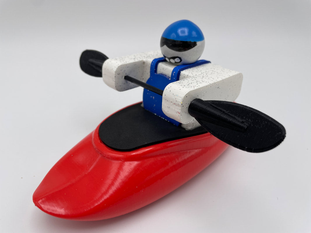 A Foamie Friends character in a blue outfit and helmet is seated in a Foamie Spud Play Boat, holding a black paddle against a white background, ready to explore river dynamics.