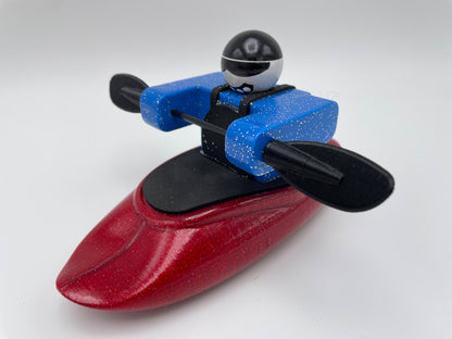 A Foamie Friends toy figure from the Foamie Spud Play Boat collection, dressed in a blue jacket and helmet, is seated in a red kayak. Designed for river dynamics enthusiasts, the figure grips black paddles against a white background.
