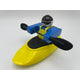 A toy figure from the Foamie Friends brand, wearing blue and black attire, skillfully navigates river dynamics while kayaking with the Foamie Spud Play Boat and holding a paddle against a white background.