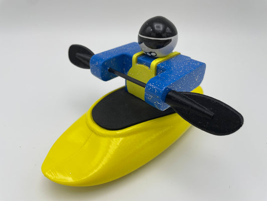 A toy figure from the Foamie Friends brand, wearing blue and black attire, skillfully navigates river dynamics while kayaking with the Foamie Spud Play Boat and holding a paddle against a white background.