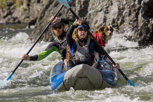 Two people in wetsuits and life jackets paddle a blue 4CRS Rental Inflatable Kayak through whitewater rapids, with a brown and white dog in a red vest sitting behind them.