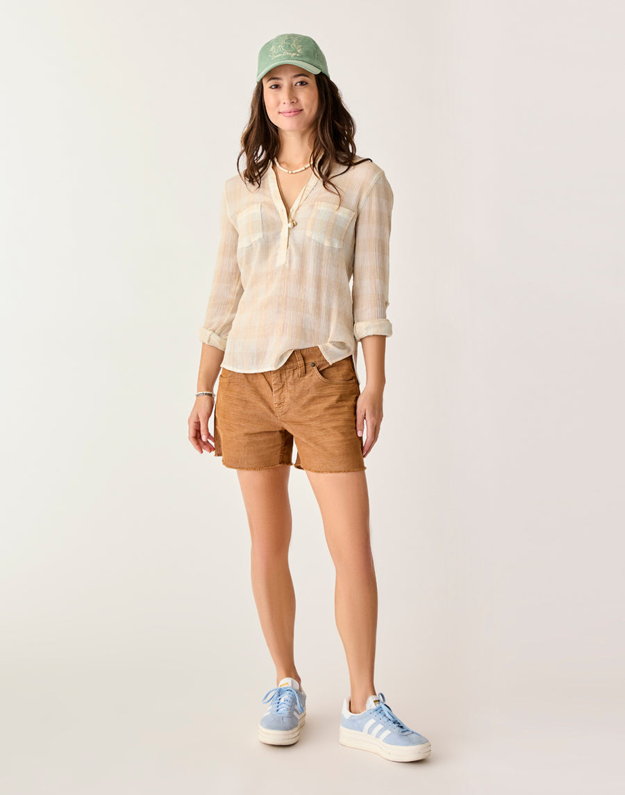 A woman faces forward in Carve's Oahu 4" Short (brown stretch cotton corduroy), a light plaid shirt, blue sneakers, and a green cap—ideal for any vacation wardrobe—set against a neutral background.