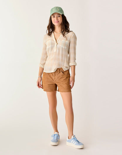 A woman faces forward in Carve's Oahu 4" Short (brown stretch cotton corduroy), a light plaid shirt, blue sneakers, and a green cap—ideal for any vacation wardrobe—set against a neutral background.