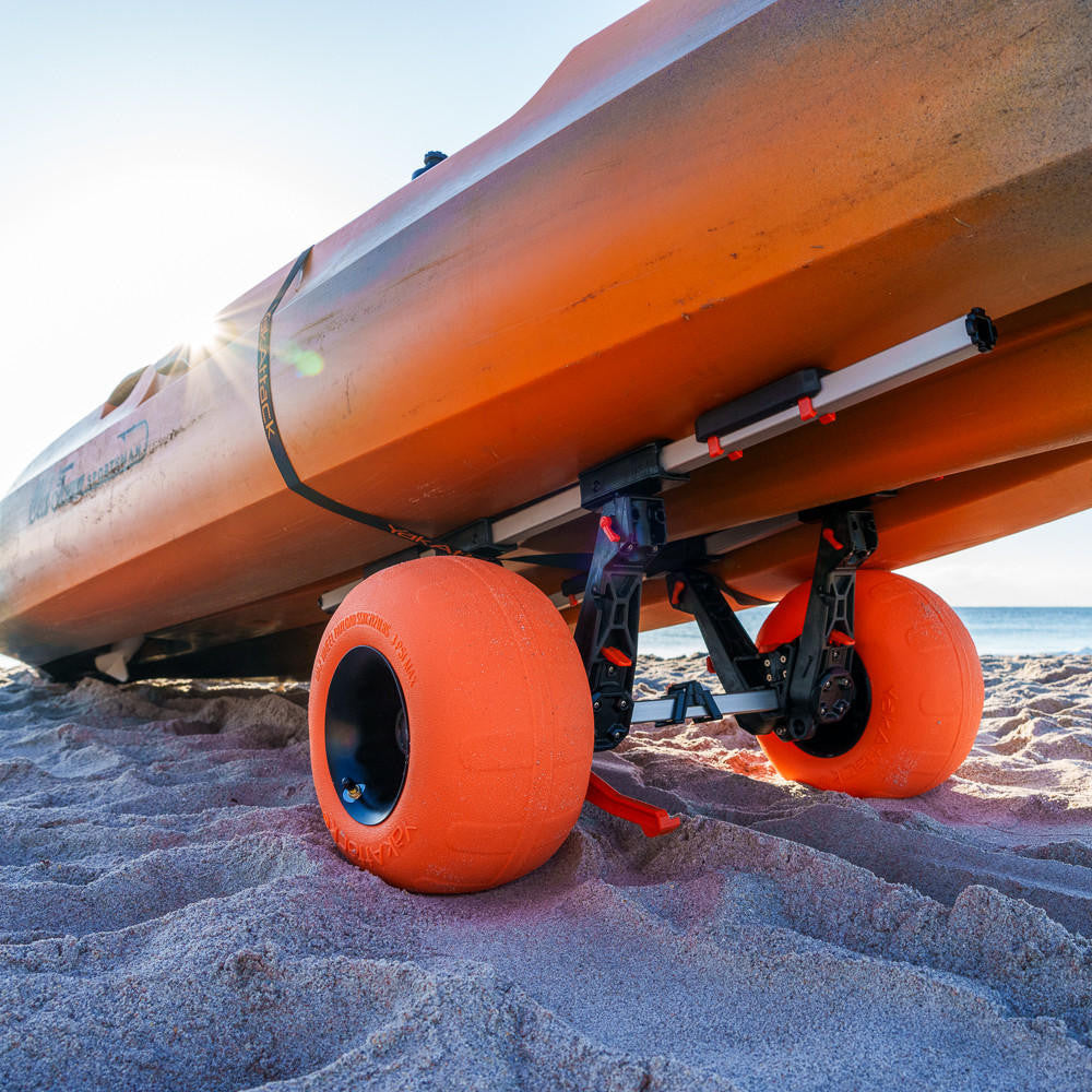 A close-up of the YakAttack TowNStow BarCart Kayak Cart with large orange sand tires, attached to a kayak on a sunny, sandy beach.