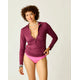 A woman smiles at the camera, wearing the Carve Cruz Rashguard over a bikini top and pink bikini bottoms, standing against a plain background.