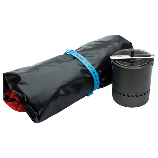 A rolled-up black and red Alpacka Mule dry bag, perfect for packraft adventures, is secured with a blue strap beside a closed silver camping pot with handles.