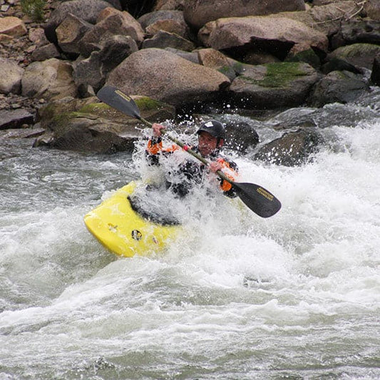 A kayaker expertly navigates Level 2 white-water rapids, skillfully maneuvering a yellow kayak with a paddle in the "Adult Level 2 Kayak Class- Sunday" by 4CRS Paddle School, against a backdrop of rugged rocks that highlights the thrill of advanced river maneuvering.