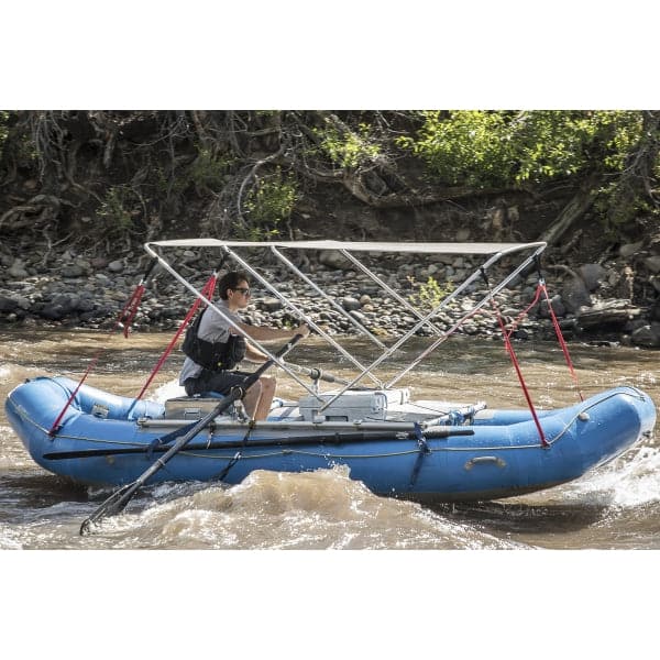 A person expertly rows a blue inflatable raft with a Coyote River Gear Raft Bimini canopy down a river flanked by rocky shores and lush trees, benefiting from the canopy's durable marine fabric for protection against the elements.
