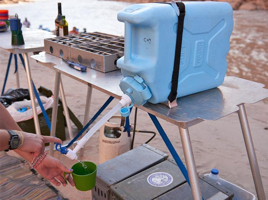 A person is filling a green cup from a blue container on the Scepter Water Jug Stand by Down River Equipment, placed on an outdoor metal table.