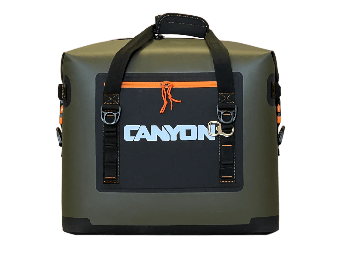 The Nomad Series Soft Cooler by Canyon Coolers features a green and black insulated design with "Canyon" text, a handle, orange waterproof zipper pocket for ice retention, and black straps with metal clasps against a plain background.