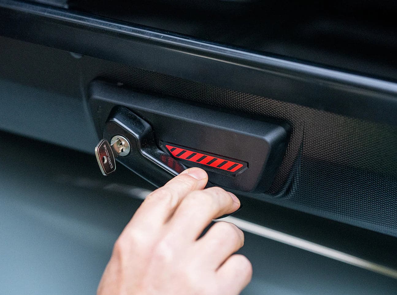 A hand turns a key in a lock featuring a design similar to the sleek Yakima Skybox NX XL cargo box, known for its black finish with a red stripe, all set against a dark surface.