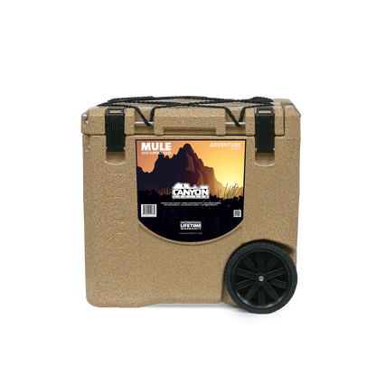 The Canyon Coolers Mule 30 Cooler in tan is a wheeled unit with hard wheels and a mountain graphic. It has two black latches, a sturdy side handle for easy transport, and a jungle bungee strap to secure essentials for adventures.