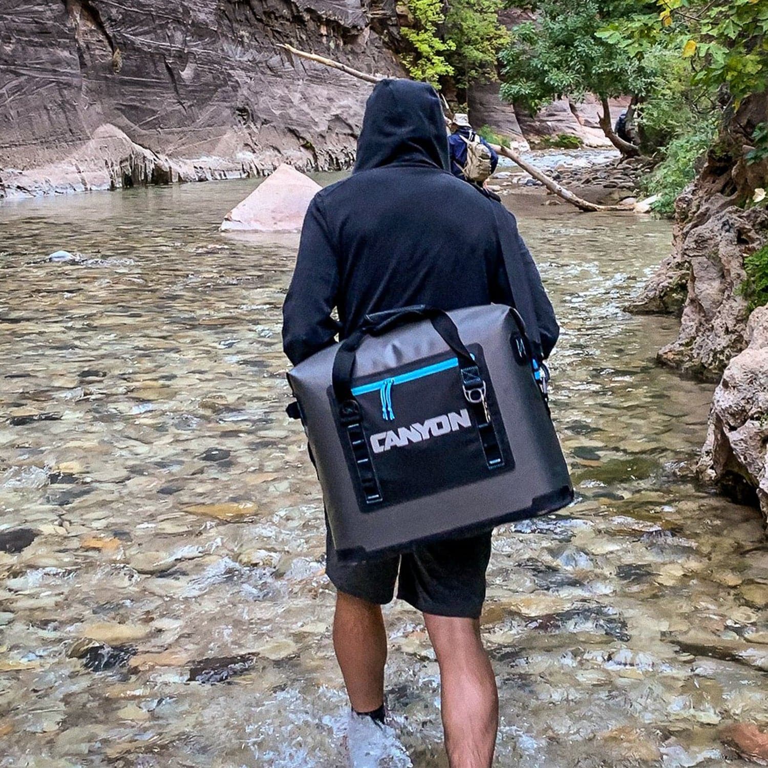 Clad in a hooded jacket, a person confidently traverses a shallow rocky river surrounded by cliffs and lush greenery, carrying a Canyon Coolers Nomad Series Soft Cooler with its waterproof zipper securely sealed.
