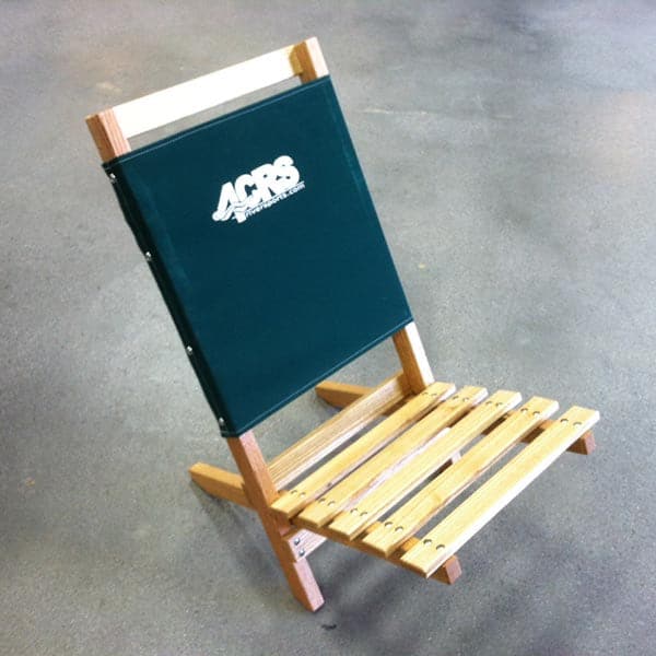 The 4CRS Lowrider Folding Chair by Coyote River Gear is a portable wooden chair with a green fabric backrest and white logo, perfect for camping. Positioned on a gray concrete floor, it features weather-resistant durability.