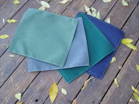 A set of fabric napkins in green, gray, teal, and blue are neatly stacked on a wooden surface with yellow leaves. Ideal for a cozy picnic using the 4CRS Lowrider Folding Chair by Coyote River Gear, they blend seamlessly into your outdoor setting.